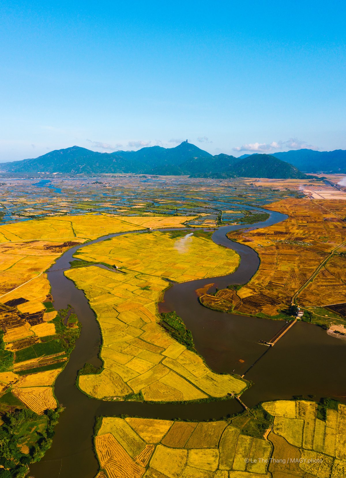 Bức ảnh chụp toàn cảnh trên không của núi Đá Bia và cánh đồng lúa vàng ở Phú Yên với dòng sông uốn lượn qua vùng nông thôn. Toàn cảnh mang lại cảm giác yên bình và rộng lớn của thiên nhiên vùng quê.