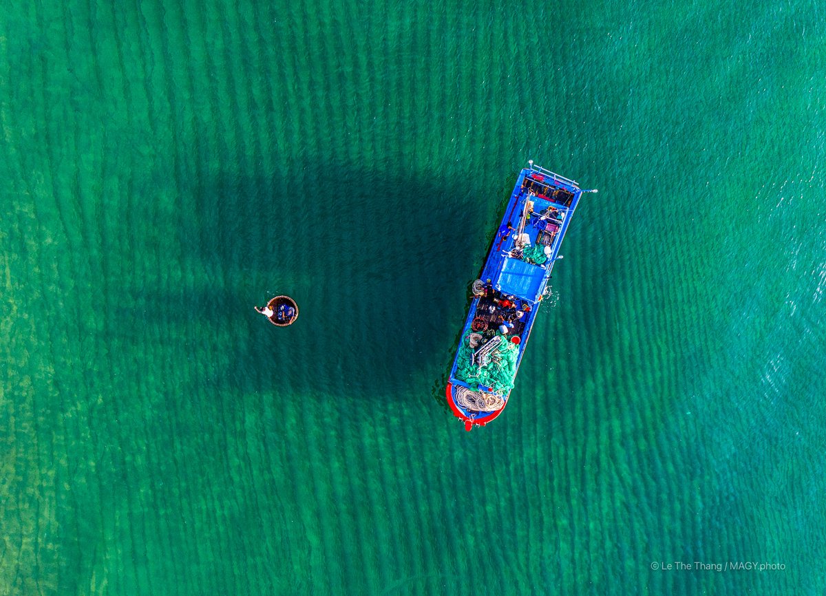 Ảnh chụp từ trên cao cho thấy một chiếc tàu cá và một thuyền thúng trên vùng biển xanh trong. Hình ảnh mang lại cảm giác yên bình và truyền tải hoạt động đánh cá truyền thống của làng chài ven biển.