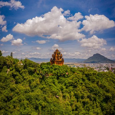 Hình ảnh tháp Nhạn cổ kính nằm trên đỉnh đồi phủ đầy cây xanh với bầu trời xanh và những đám mây trắng phía trên. Cảnh quan tạo cảm giác yên bình và hài hoà giữa thiên nhiên và kiến trúc lịch sử.