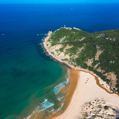 Hình ảnh chụp từ trên cao cho thấy mũi đất với đèn biển nằm trên đỉnh đồi, bao quanh bởi biển xanh và bãi cát vàng. Cảnh quan mang lại cảm giác yên bình và tĩnh lặng giữa thiên nhiên rộng lớn.