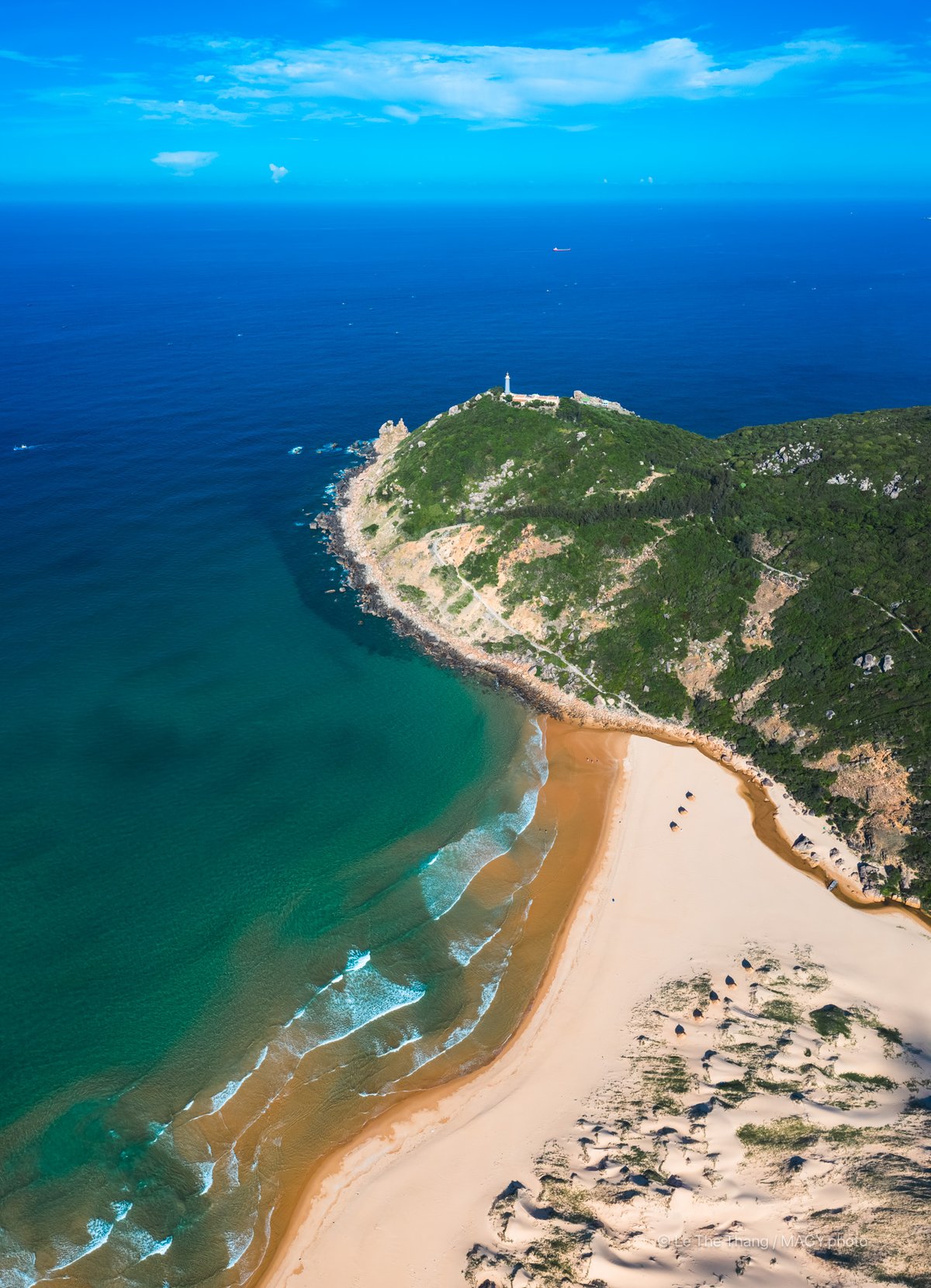 Hình ảnh chụp từ trên cao cho thấy mũi đất với đèn biển nằm trên đỉnh đồi, bao quanh bởi biển xanh và bãi cát vàng. Cảnh quan mang lại cảm giác yên bình và tĩnh lặng giữa thiên nhiên rộng lớn.