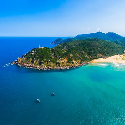 Ảnh chụp toàn cảnh trên không của Mũi Điện, Phú Yên, với bãi biển cát vàng, núi xanh và biển xanh trong ánh sáng tự nhiên. Cảnh quan tạo cảm giác yên bình và tươi mát, thích hợp cho du lịch và nghỉ dưỡng.