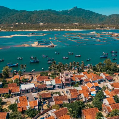 Ảnh chụp toàn cảnh trên không của một làng ven biển với nhiều tàu cá neo đậu trên biển xanh, phía sau là dãy núi xanh mướt. Cảnh vật mang lại cảm giác yên bình và tĩnh lặng của một vùng ven biển miền Trung Việt Nam.