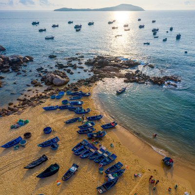 Ảnh chụp toàn cảnh trên cao của bãi biển với nhiều tàu cá và thuyền nhỏ trên cát và biển. Cảnh biển yên bình với ánh sáng tự nhiên nhẹ nhàng tạo cảm giác thư thái và thanh bình.