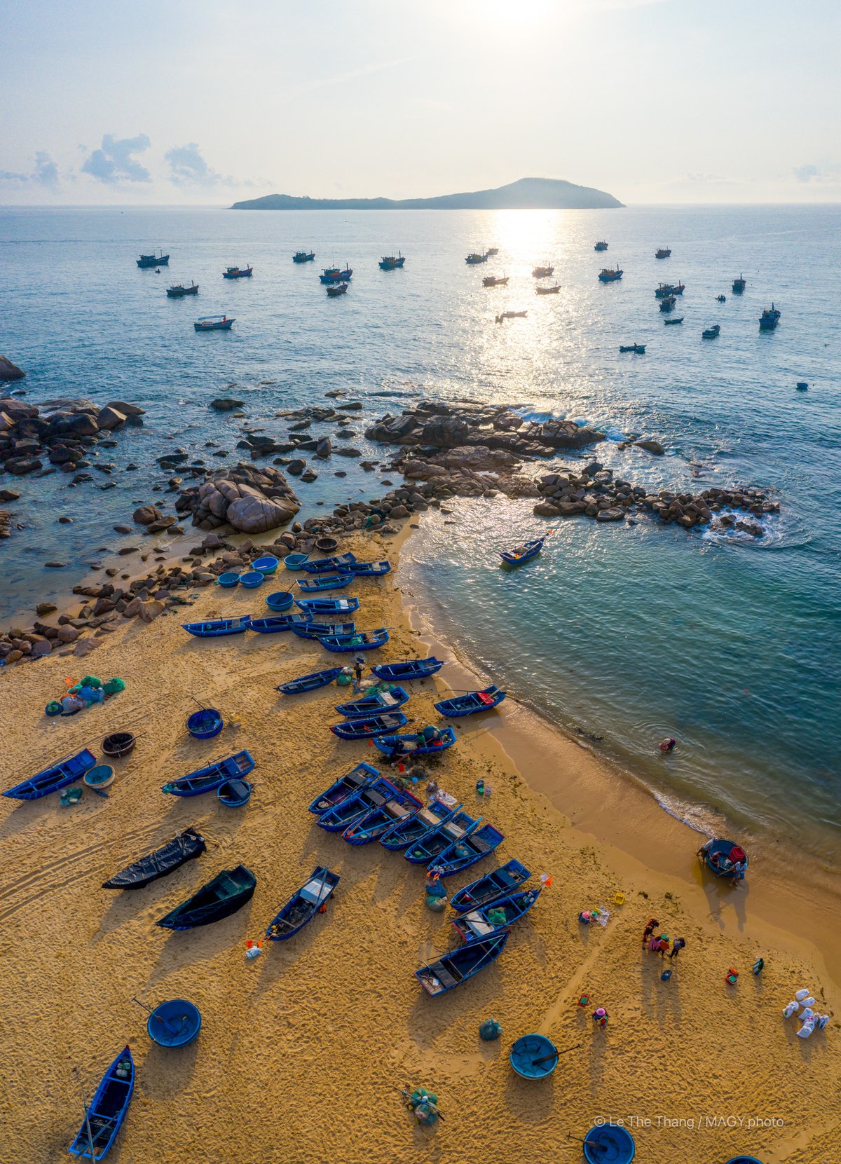 Ảnh chụp toàn cảnh trên cao của bãi biển với nhiều tàu cá và thuyền nhỏ trên cát và biển. Cảnh biển yên bình với ánh sáng tự nhiên nhẹ nhàng tạo cảm giác thư thái và thanh bình.