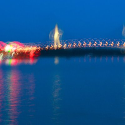 Bức ảnh chụp cầu Cần Thơ vào giờ xanh với ánh sáng đèn chiếu sáng mờ ảo phản chiếu trên mặt nước sông. Tổng thể tạo cảm giác yên bình và huyền ảo trong không gian buổi tối.