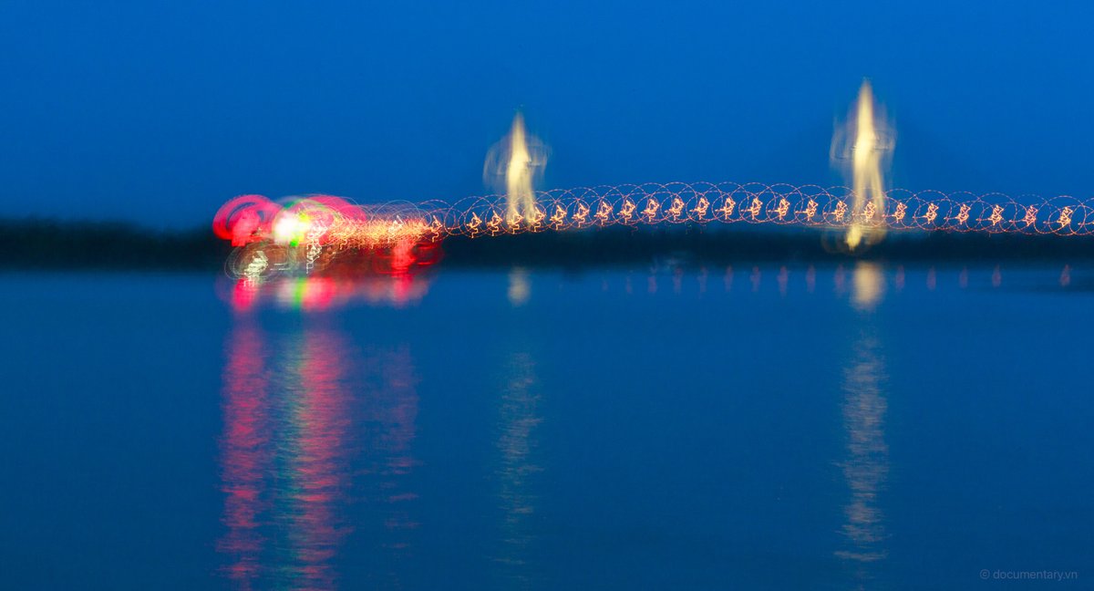 Bức ảnh chụp cầu Cần Thơ vào giờ xanh với ánh sáng đèn chiếu sáng mờ ảo phản chiếu trên mặt nước sông. Tổng thể tạo cảm giác yên bình và huyền ảo trong không gian buổi tối.