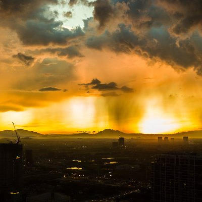 Bức ảnh chụp cảnh hoàng hôn tại một thành phố với mưa rơi và những đám mây đen trên bầu trời. Cảnh vật tạo cảm giác mạnh mẽ và đầy kịch tính với ánh sáng vàng rực rỡ xuyên qua mưa.