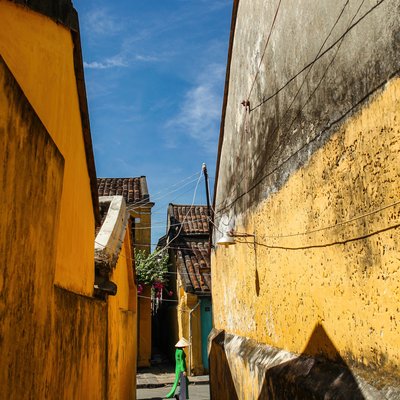 Bức ảnh chụp một người phụ nữ mặc áo dài xanh và đội nón lá đi trong một ngõ hẹp với các bức tường vàng cổ kính dưới ánh sáng tự nhiên. Cảnh vật mang lại cảm giác yên bình và truyền thống đặc trưng của phố cổ Hội An.