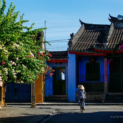 Bức ảnh chụp một người phụ nữ đội nón lá đang đạp xe trên con phố cổ với kiến trúc truyền thống và hoa rực rỡ hai bên đường dưới ánh sáng tự nhiên. Cảnh vật mang lại cảm giác yên bình và hoài cổ về cuộc sống thường nhật ở Hội An.