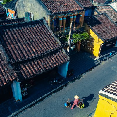 Bức ảnh chụp một người bán hàng truyền thống trên đường phố phố cổ Hội An với mái ngói đỏ và tường vàng đặc trưng. Cảnh vật mang lại cảm giác yên bình và truyền thống của một thị trấn cổ kính.