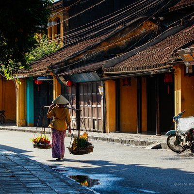Bức ảnh chụp một người phụ nữ đội nón lá đang bán hoa trên phố cổ Hội An với ánh sáng tự nhiên chiếu sáng các toà nhà vàng truyền thống. Cảnh vật mang lại cảm giác yên bình và hoài cổ về cuộc sống thường nhật tại phố cổ.