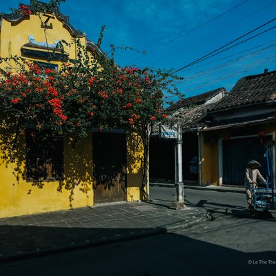 Bức ảnh chụp một con phố cổ với các toà nhà màu vàng đặc trưng và hoa đỏ rực rỡ dưới ánh sáng tự nhiên buổi sáng. Cảnh vật mang lại cảm giác yên bình và bình dị của cuộc sống đô thị truyền thống.