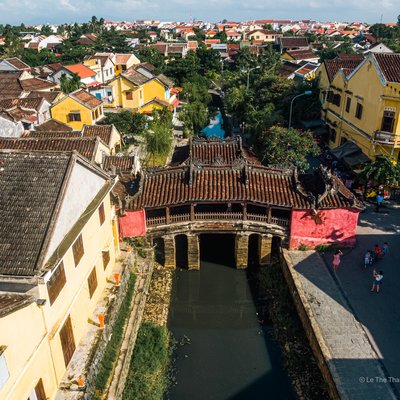 Hình ảnh chụp toàn cảnh cầu cổ Chùa Cầu tại Hội An với các ngôi nhà vàng truyền thống và dòng sông chảy qua. Cảnh quan mang lại cảm giác yên bình và cổ kính dưới ánh sáng tự nhiên của bầu trời trong xanh.