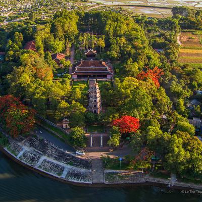Ảnh chụp toàn cảnh trên không của chùa Thiên Mụ tại Huế, nằm bên bờ sông với kiến trúc cổ và nhiều cây xanh bao quanh. Cảnh vật tạo cảm giác yên bình và thanh tịnh giữa thiên nhiên và lịch sử.