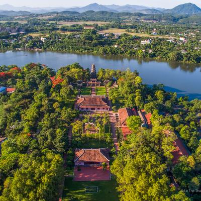 Ảnh toàn cảnh trên không của chùa Thiên Mụ nằm bên bờ sông trong khu vực nhiều cây xanh và địa hình bằng phẳng. Khung cảnh mang lại cảm giác yên bình và tĩnh lặng giữa thiên nhiên và kiến trúc truyền thống.