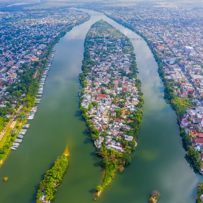 Ảnh chụp từ trên không cho thấy sông Hương với đảo Cồn Hến ở giữa, bao quanh bởi khu đô thị và cây xanh. Cảnh quan mang lại cảm giác yên bình và tĩnh lặng của một thành phố ven sông.