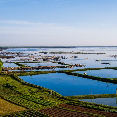 Bức ảnh chụp cảnh bình minh trên phá Tam Giang với các thuyền đánh cá và cánh đồng nông nghiệp xanh mướt. Toàn cảnh tạo cảm giác yên bình và tĩnh lặng của vùng nông thôn miền Trung Việt Nam.