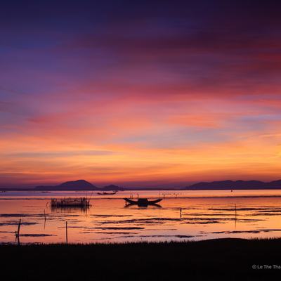 Bức ảnh chụp bình minh tại đầm Cầu Hai với ánh sáng tự nhiên màu cam rực rỡ phản chiếu trên mặt nước phẳng lặng. Khung cảnh núi non xa xa và thuyền nhỏ tạo nên cảm giác bình yên và thanh tĩnh.