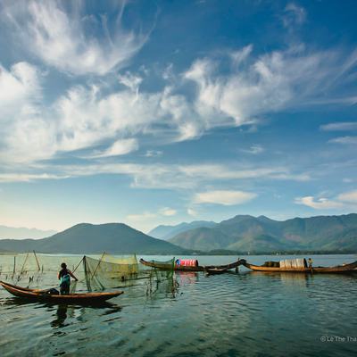 Bức ảnh chụp cảnh ngư dân đang chèo thuyền và đánh cá trên đầm phá Tam Giang với núi non phía xa và bầu trời trong xanh lúc bình minh. Cảnh vật mang lại cảm giác yên bình và tĩnh lặng của cuộc sống miền quê ven đầm phá.