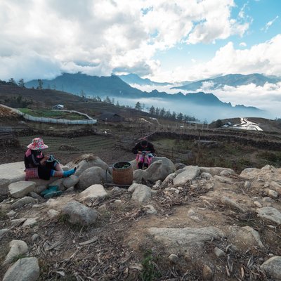 Bức ảnh chụp cảnh núi non và cao nguyên với những đám mây bao phủ, có hai người đang ngồi nghỉ trên đá. Cảnh vật tạo cảm giác yên bình và tĩnh lặng giữa thiên nhiên rộng lớn.