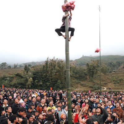 Bức ảnh chụp một lễ hội truyền thống tại vùng nông thôn với nhiều người tham gia, trong đó một người đang leo lên cột tre để lấy phần thưởng. Không khí lễ hội sôi động và đầy màu sắc, thể hiện sự đoàn kết và vui tươi của cộng đồng.