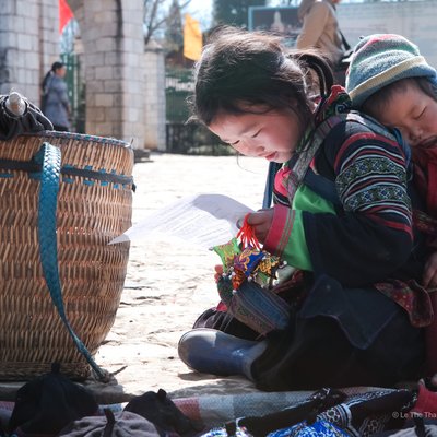 Bức ảnh chụp một bé gái dân tộc đang ngồi học chữ ngoài trời với em bé được địu trên lưng. Cảnh tượng toát lên sự bình dị và tình cảm gia đình trong cuộc sống thường nhật.