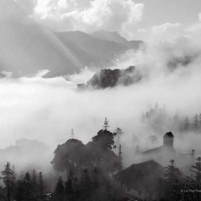 Bức ảnh đen trắng thể hiện cảnh núi non và làng mạc ở Sa Pa với sương mù bao phủ và ánh sáng tự nhiên dịu nhẹ. Toàn cảnh tạo cảm giác huyền bí và yên bình, như một thị trấn ẩn mình trong mây.