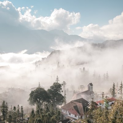 Hình ảnh thể hiện cảnh quan thị trấn Sa Pa trong sương mù với ánh sáng tự nhiên và bầu trời xanh. Toàn cảnh tạo cảm giác yên bình và huyền ảo của vùng núi Tây Bắc Việt Nam.