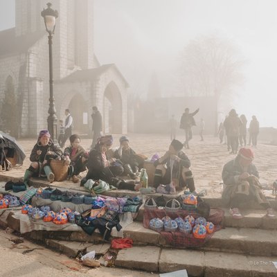 Bức ảnh chụp một nhóm người dân tộc ngồi trên bậc thềm ngoài trời trong sương mù nhẹ, bán các sản phẩm thủ công mỹ nghệ truyền thống. Không khí yên bình và mang đậm nét văn hoá đặc trưng của vùng cao Tây Bắc Việt Nam.