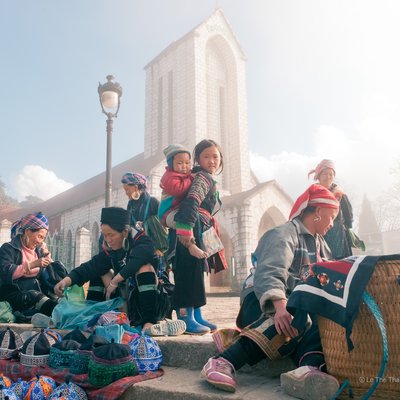 Hình ảnh chợ ngoài trời tại thị trấn Sa Pa vào buổi sáng với ánh sáng tự nhiên dịu nhẹ và bầu trời xanh. Cảnh tượng mang lại cảm giác bình yên và truyền thống với những người dân tộc mặc trang phục đặc trưng đang giao thương.