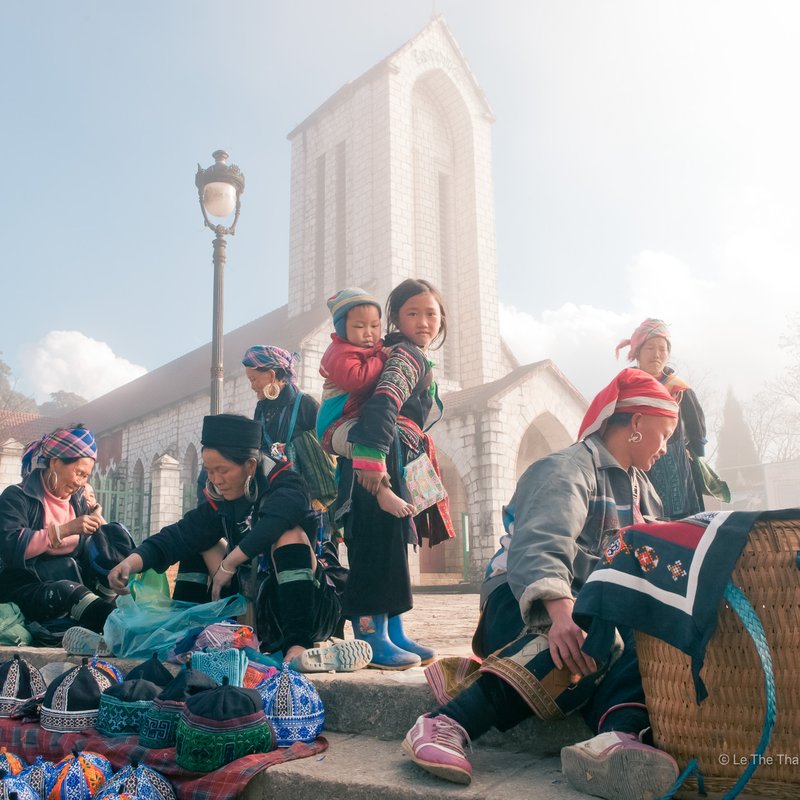 Hình ảnh chợ ngoài trời tại thị trấn Sa Pa vào buổi sáng với ánh sáng tự nhiên dịu nhẹ và bầu trời xanh. Cảnh tượng mang lại cảm giác bình yên và truyền thống với những người dân tộc mặc trang phục đặc trưng đang giao thương.