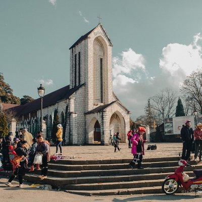 Bức ảnh chụp một nhà thờ bằng đá ở thị trấn Sapa với nhiều người dân địa phương đang tụ tập và giao tiếp xung quanh. Không khí trong ảnh mang lại cảm giác bình yên và chân thực về cuộc sống thường nhật của người dân nơi đây.