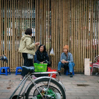 Bức ảnh chụp một quán cóc nhỏ trên vỉa hè với vài người đang ngồi và trò chuyện bên cạnh một bức tường tre. Không gian tạo cảm giác thân thiện và bình dị, thể hiện nét văn hoá đường phố đặc trưng của Hà Nội.