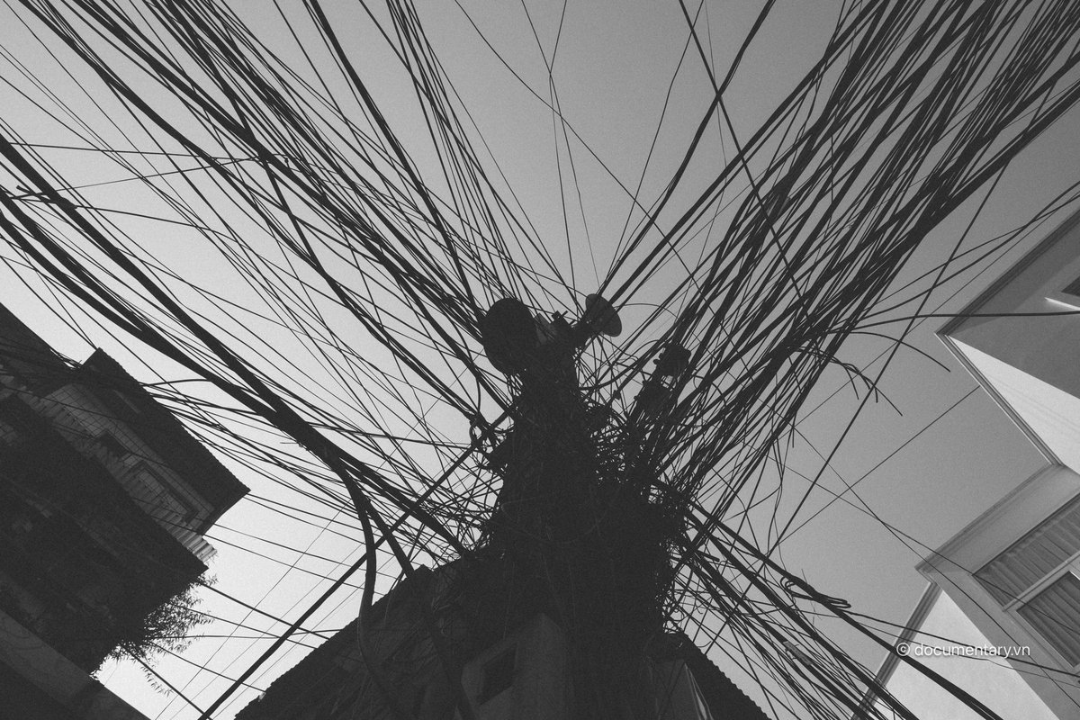 Hình ảnh chụp một cột điện với rất nhiều dây cáp quang và dây điện chằng chịt. Bức ảnh tạo cảm giác hỗn độn và phức tạp trong hệ thống dây điện đô thị.