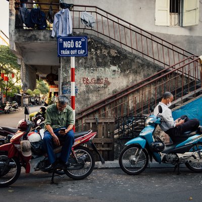 Bức ảnh chụp hai người đàn ông ngồi trên xe máy đậu bên lề đường trong một con ngõ ở Hà Nội. Không khí yên bình và tĩnh lặng tạo cảm giác thư thái và bình dị của cuộc sống hàng ngày.