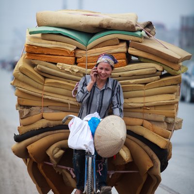 Một người phụ nữ đang đi xe đạp chở một khối lượng lớn đệm mút trên đường phố Hà Nội và đang nói chuyện điện thoại. Hình ảnh thể hiện sự bận rộn và đa nhiệm trong cuộc sống hàng ngày ở thành phố.
