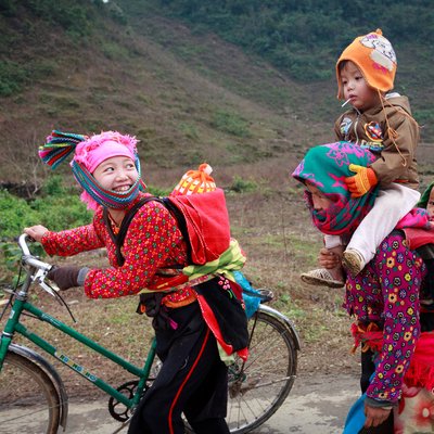 Bức ảnh chụp cảnh hai phụ nữ dân tộc thiểu số đang đưa trẻ em đi học ở vùng núi. Không khí trong ảnh thể hiện sự ấm áp và vui tươi giữa cảnh trời lạnh và khung cảnh thiên nhiên xung quanh.