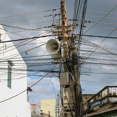 Bức ảnh chụp một cột điện với nhiều dây cáp viễn thông và dây điện chằng chịt ở một khu phố đô thị. Không khí trong ảnh mang lại cảm giác yên bình với ánh sáng mềm và bầu trời nhiều mây.