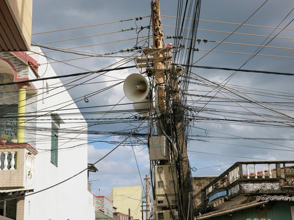 Bức ảnh chụp một cột điện với nhiều dây cáp viễn thông và dây điện chằng chịt ở một khu phố đô thị. Không khí trong ảnh mang lại cảm giác yên bình với ánh sáng mềm và bầu trời nhiều mây.