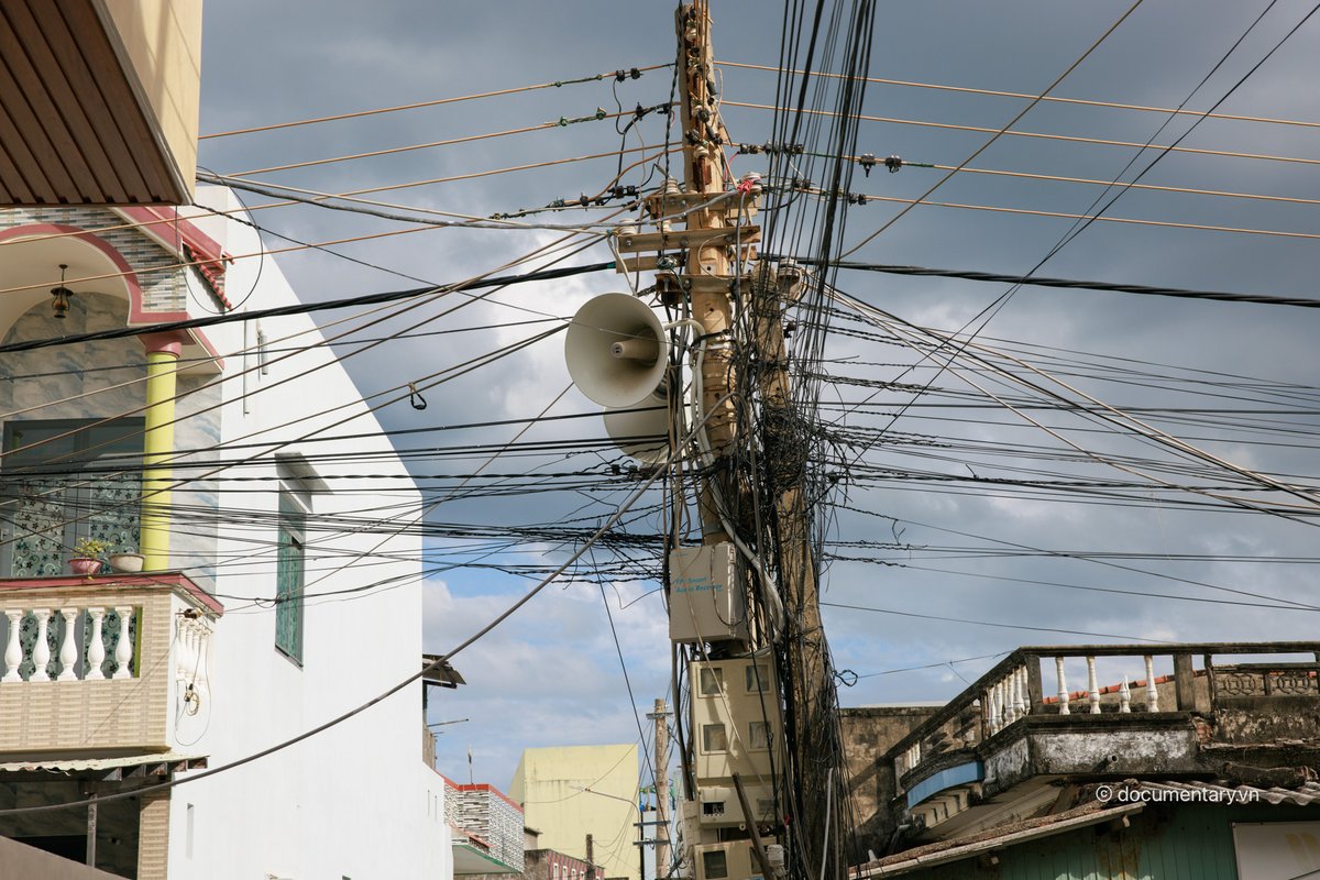Bức ảnh chụp một cột điện với nhiều dây cáp viễn thông và dây điện chằng chịt ở một khu phố đô thị. Không khí trong ảnh mang lại cảm giác yên bình với ánh sáng mềm và bầu trời nhiều mây.