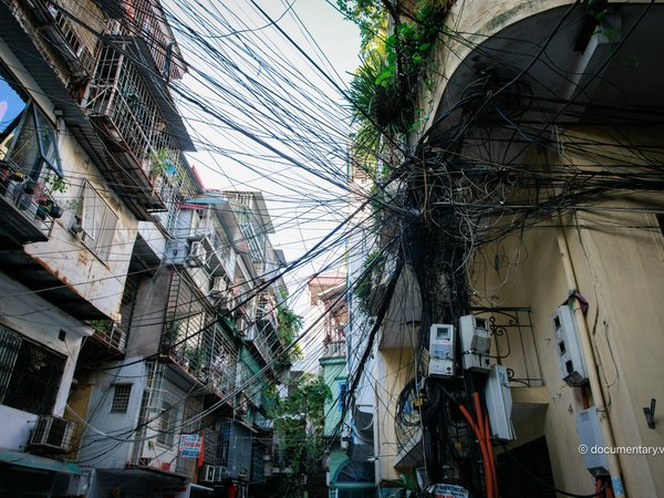 Bức ảnh chụp cảnh một con phố nhỏ ở Hà Nội với nhiều dây điện chằng chịt và các toà nhà cũ kỹ. Không khí yên bình và chân thực thể hiện cuộc sống thường nhật của thành phố.