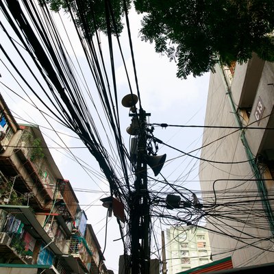 Bức ảnh chụp một khu nhà tập thể cũ với nhiều dây điện chằng chịt và các loa phường gắn trên cột điện. Hình ảnh thể hiện một phần đời sống đô thị thường nhật với cảm giác bình dị và quen thuộc.
