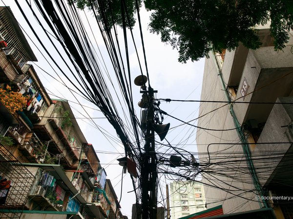Bức ảnh chụp một khu nhà tập thể cũ với nhiều dây điện chằng chịt và các loa phường gắn trên cột điện. Hình ảnh thể hiện một phần đời sống đô thị thường nhật với cảm giác bình dị và quen thuộc.