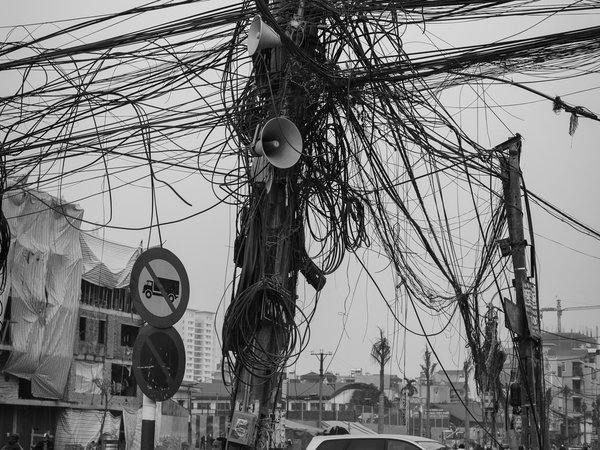 Bức ảnh chụp một cột điện với rất nhiều dây điện rối rắm ở đầu con đường mới mở rộng tại một khu vực đô thị. Cảnh vật thể hiện sự nhộn nhịp và phức tạp của giao thông cùng các phương tiện di chuyển xung quanh.