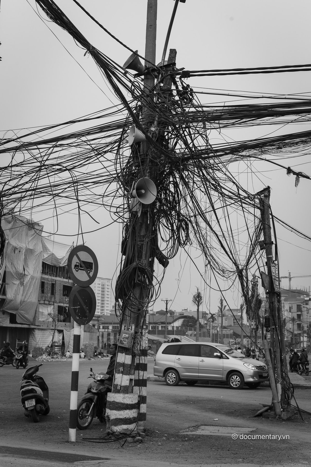 Bức ảnh chụp một cột điện với rất nhiều dây điện rối rắm ở đầu con đường mới mở rộng tại một khu vực đô thị. Cảnh vật thể hiện sự nhộn nhịp và phức tạp của giao thông cùng các phương tiện di chuyển xung quanh.
