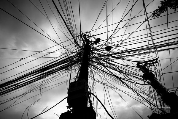 Bức ảnh chụp các dây điện chằng chịt trên cột điện trong một khu vực đô thị. Hình ảnh tạo cảm giác hỗn độn và phức tạp của hệ thống điện trong thành phố.