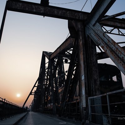 Hình ảnh chụp cầu Long Biên tại Hà Nội vào lúc hoàng hôn với ánh sáng dịu nhẹ. Cảnh vật mang lại cảm giác yên bình và tĩnh lặng, thể hiện sự kết hợp giữa kiến trúc công nghiệp và thiên nhiên.