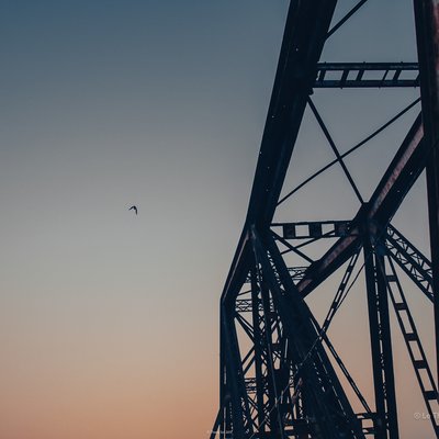 Hình ảnh chụp một phần của cầu Long Biên với bầu trời hoàng hôn và một con chim bay. Cảnh vật mang lại cảm giác yên bình và tĩnh lặng trong không gian đô thị.
