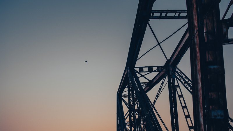 Hình ảnh chụp một phần của cầu Long Biên với bầu trời hoàng hôn và một con chim bay. Cảnh vật mang lại cảm giác yên bình và tĩnh lặng trong không gian đô thị.